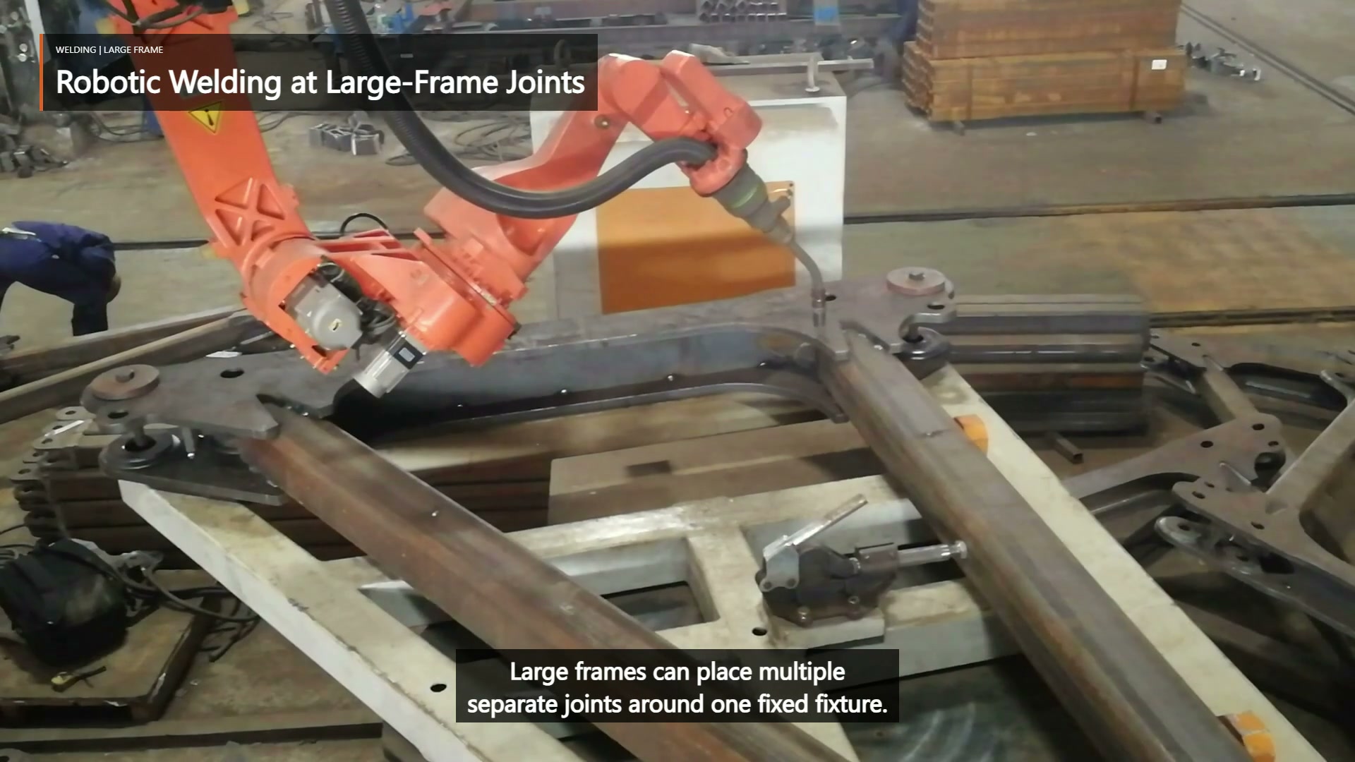 An orange robot and welding torch work beside a large fixed tubular frame in a steel fixture.