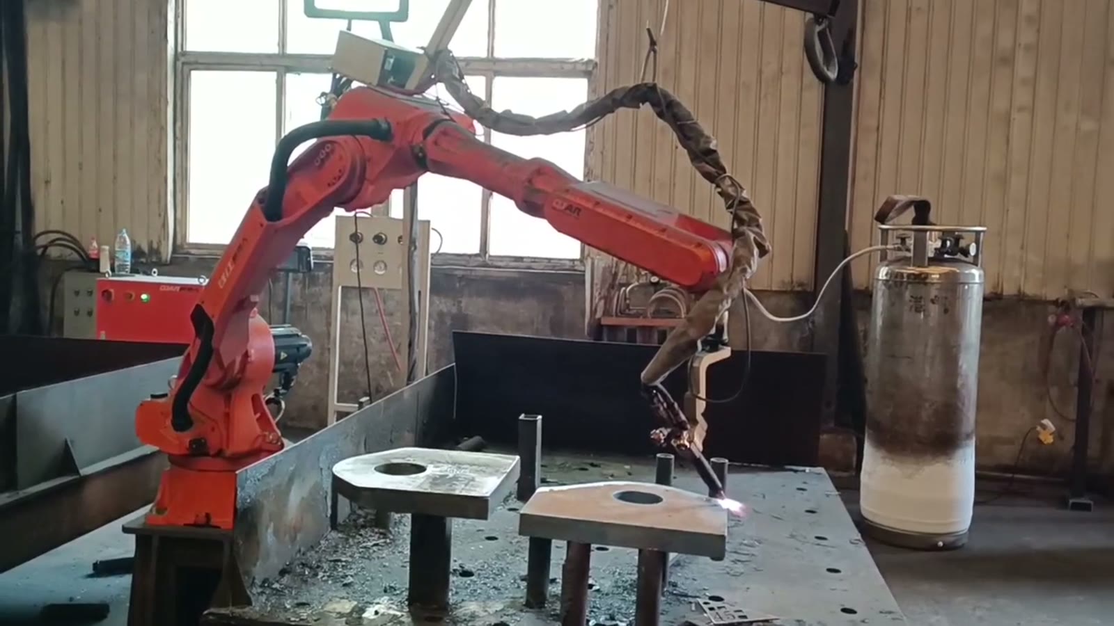 Red six-axis robot with a flame cutting torch beside two thick steel plates with round holes resting on support posts