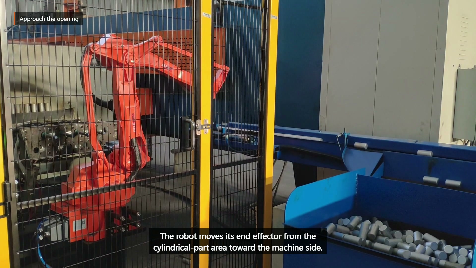 The orange robot moves toward the blue enclosed equipment beside the cylindrical-part area.