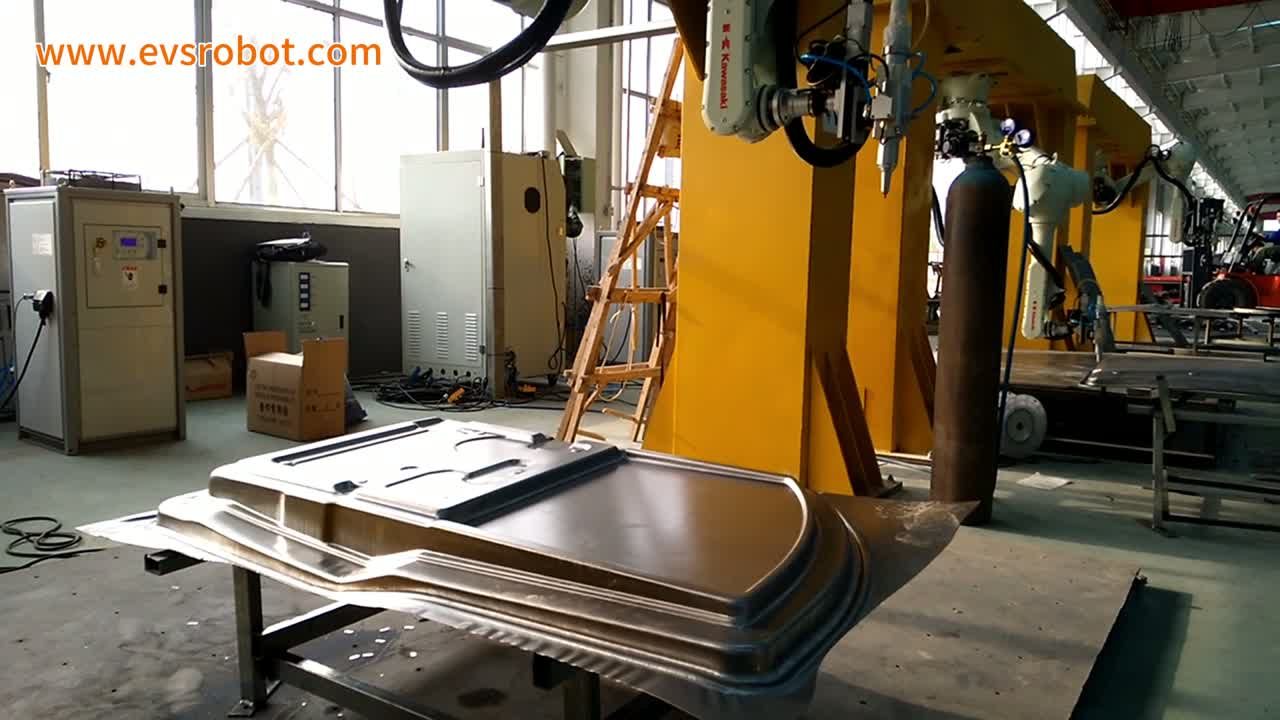 A robot arm reaches down over a large stamped panel on a square-tube stand