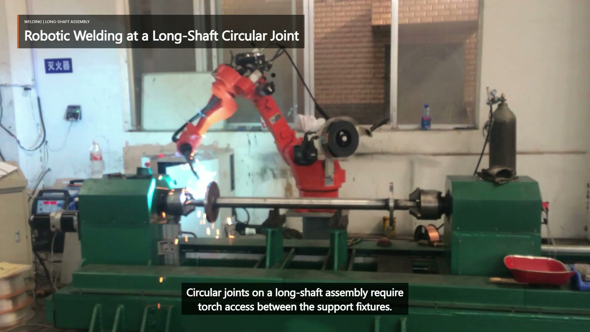 An orange robot welds beside a long horizontal shaft assembly held by green supports.