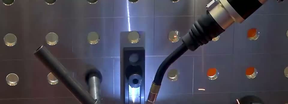Processed close-up of a welding torch working on an angled tubular joint over a fixture table