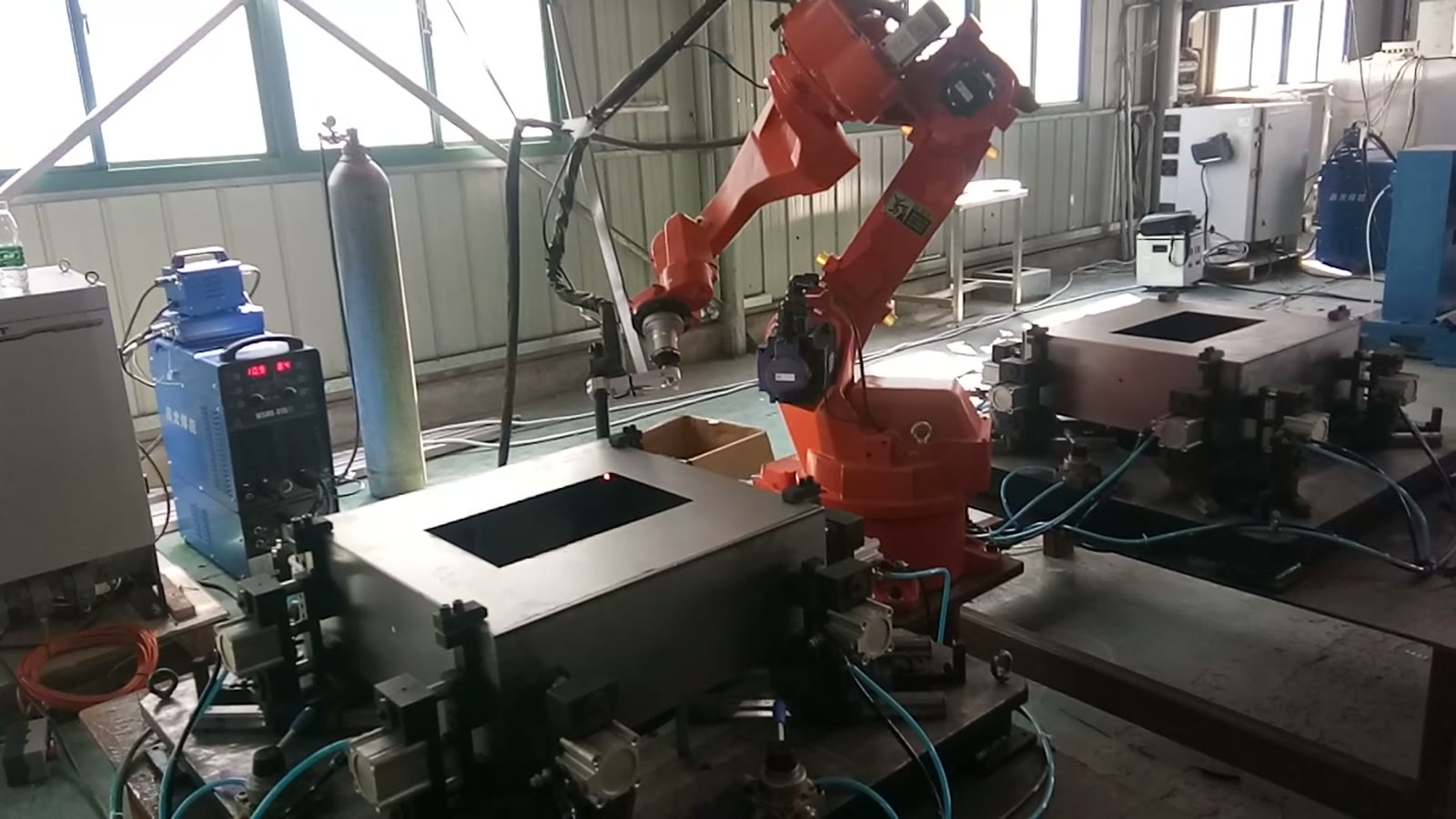 Orange six-axis welding robot between two fixture stations, each holding a square sheet-metal box