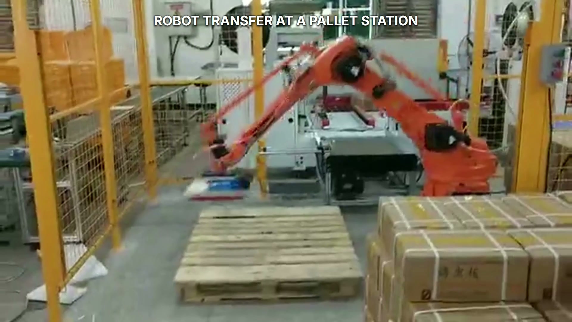 An orange industrial robot works between a rear supply station and a wooden pallet area.