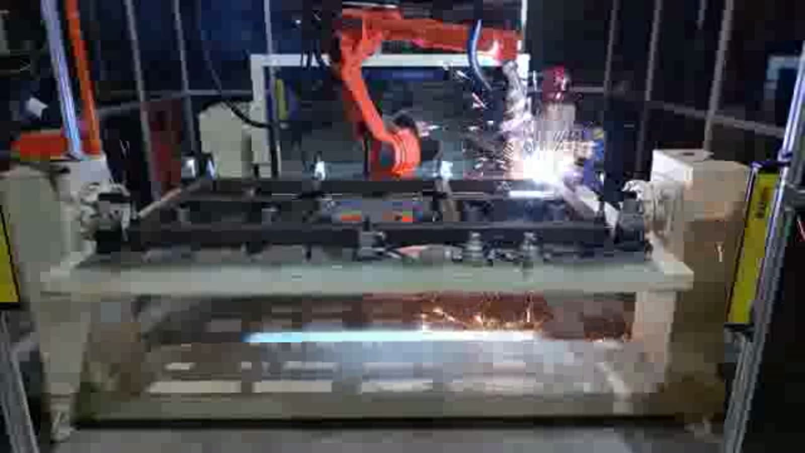 Orange six-axis welding robot behind a rectangular steel tube frame lying on a long fixture between two end stands