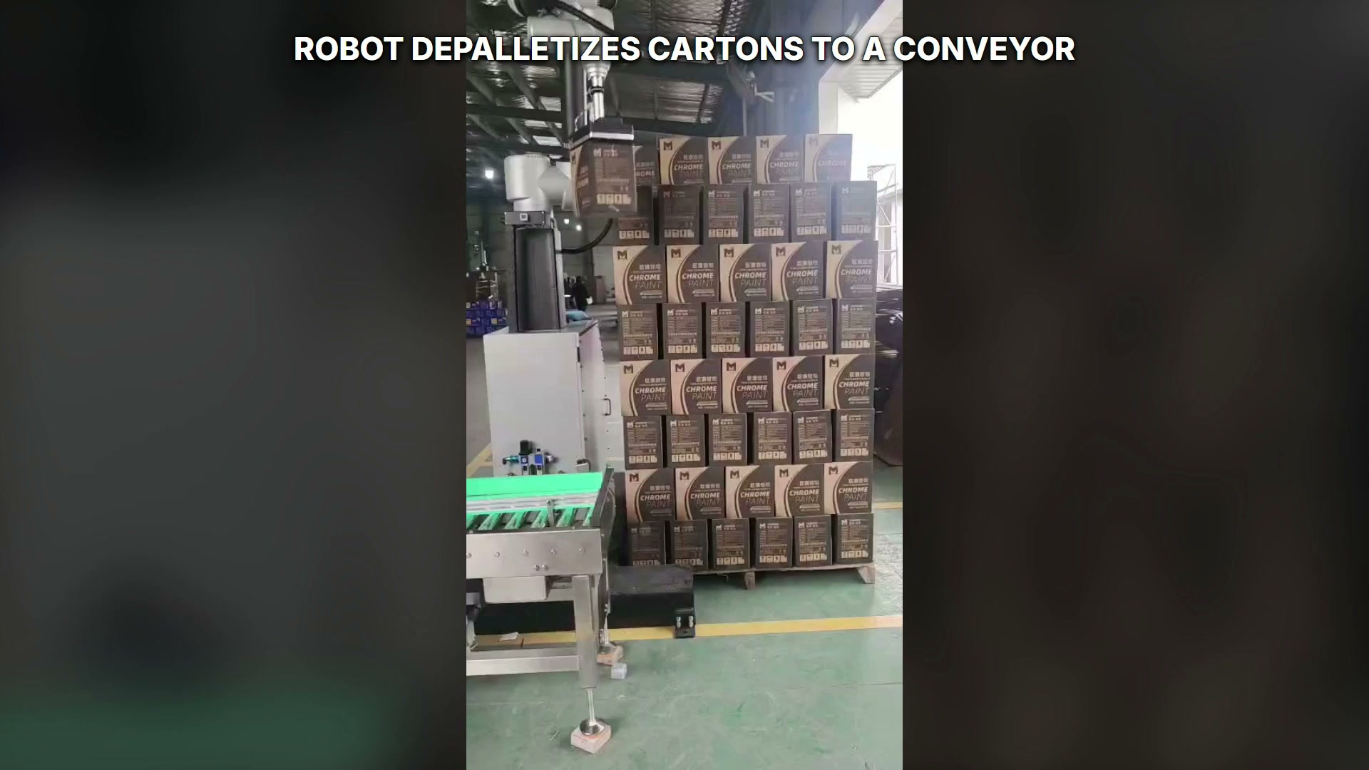 The robot holds one carton between the right-hand stack and the green conveyor.